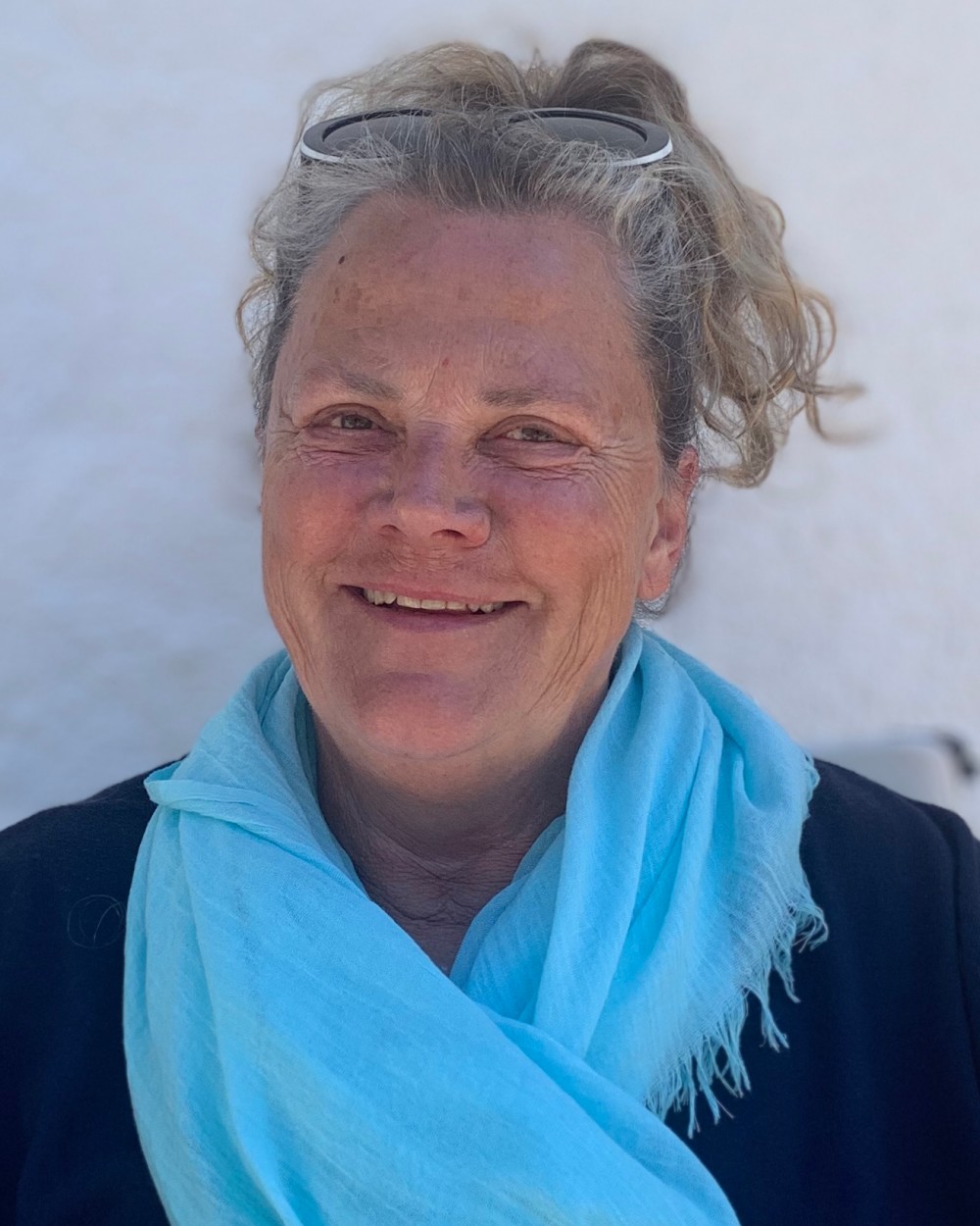 Porträt einer lächelnden Frau mit grauen, lockigen Haaren, die eine Sonnenbrille auf dem Kopf und einen leuchtend blauen Schal trägt. Der Hintergrund ist eine neutrale, helle Wand.