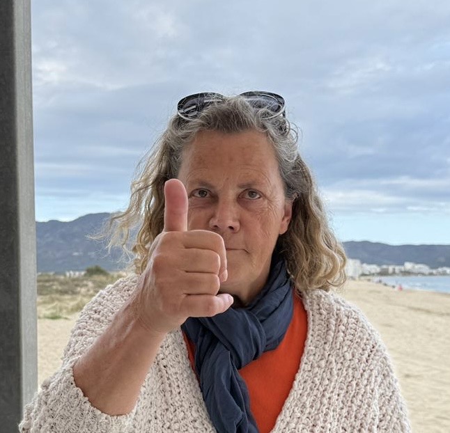 Ältere Frau am Strand blickt in die Kamera und zeigt Daumen hoch. Sie trägt Schal, Strickjacke und Sonnenbrille auf dem Kopf.